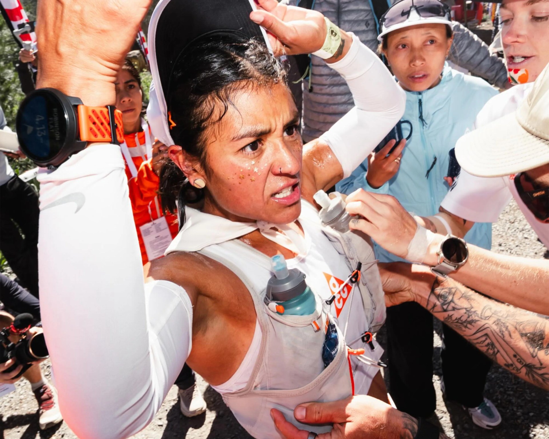 Athlete in white sportswear and hydration vest raising arms, surrounded by people, one with tattooed arm.