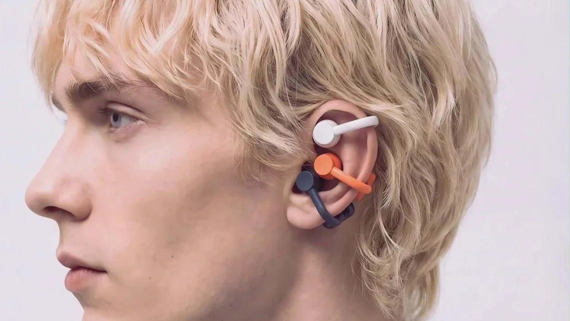 Side profile of a person with blonde hair wearing three wireless earbuds in white, orange, and black on one ear.