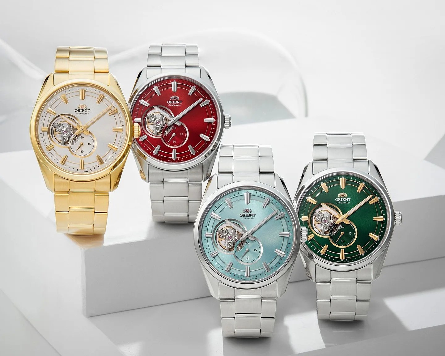 Four Orient automatic wristwatches with metal bands in gold, silver with red dial, silver with light blue dial, and silver with green dial on a white surface.