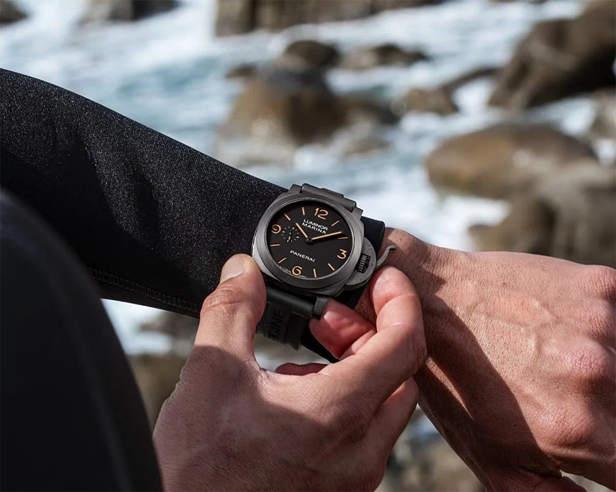 Black Panerai Luminor Marina wristwatch with orange numerals worn on a wrist near a rocky shoreline.