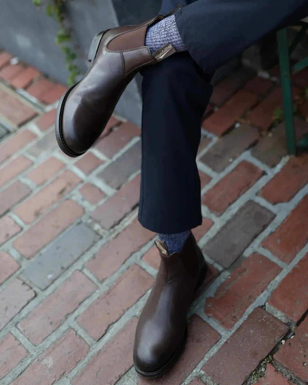 Brown leather Chelsea boots worn with blue socks and dark pants on a brick pavement.