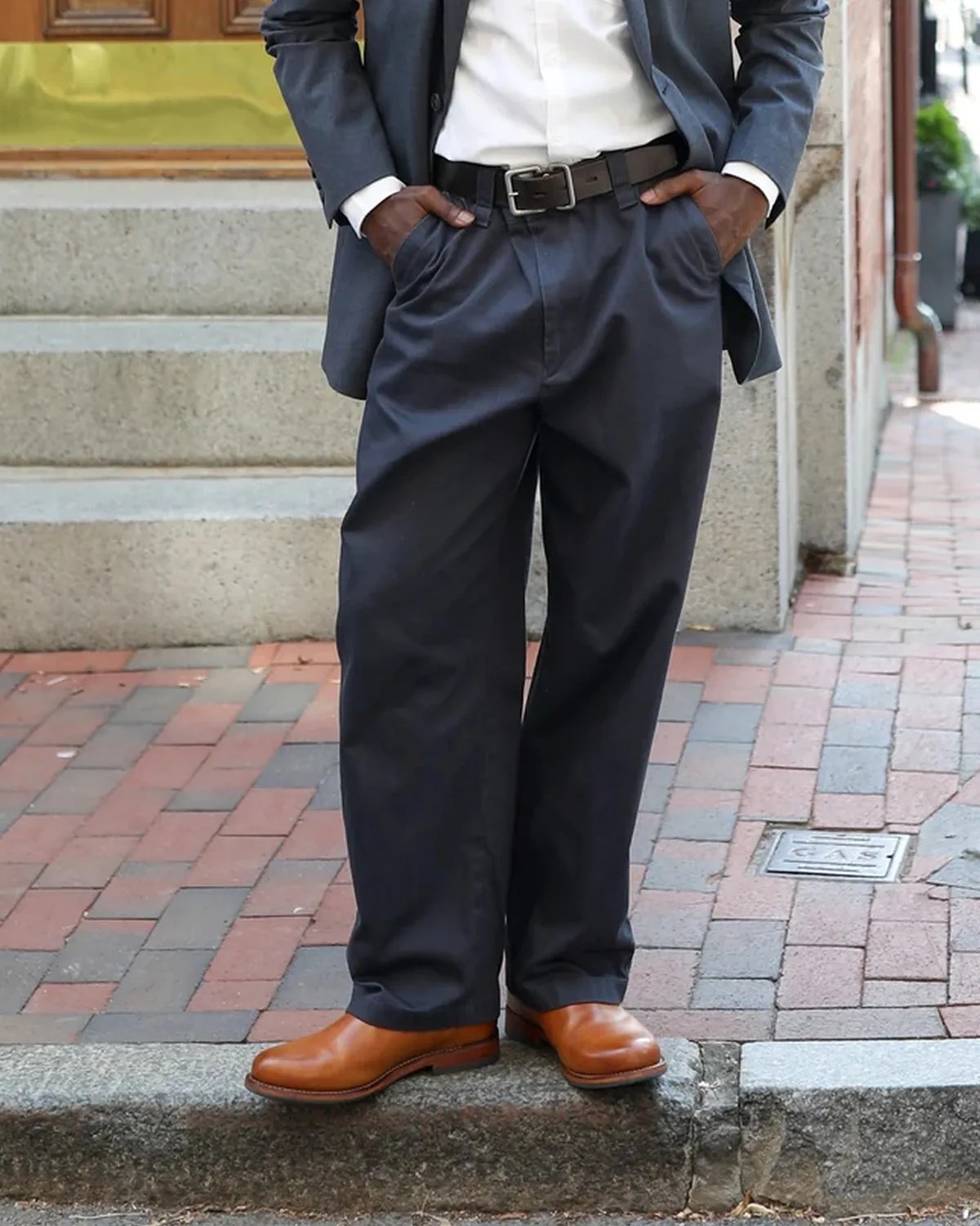 Man wearing loose dark navy trousers, brown leather shoes, black belt, white shirt, and gray blazer standing on a stone step.