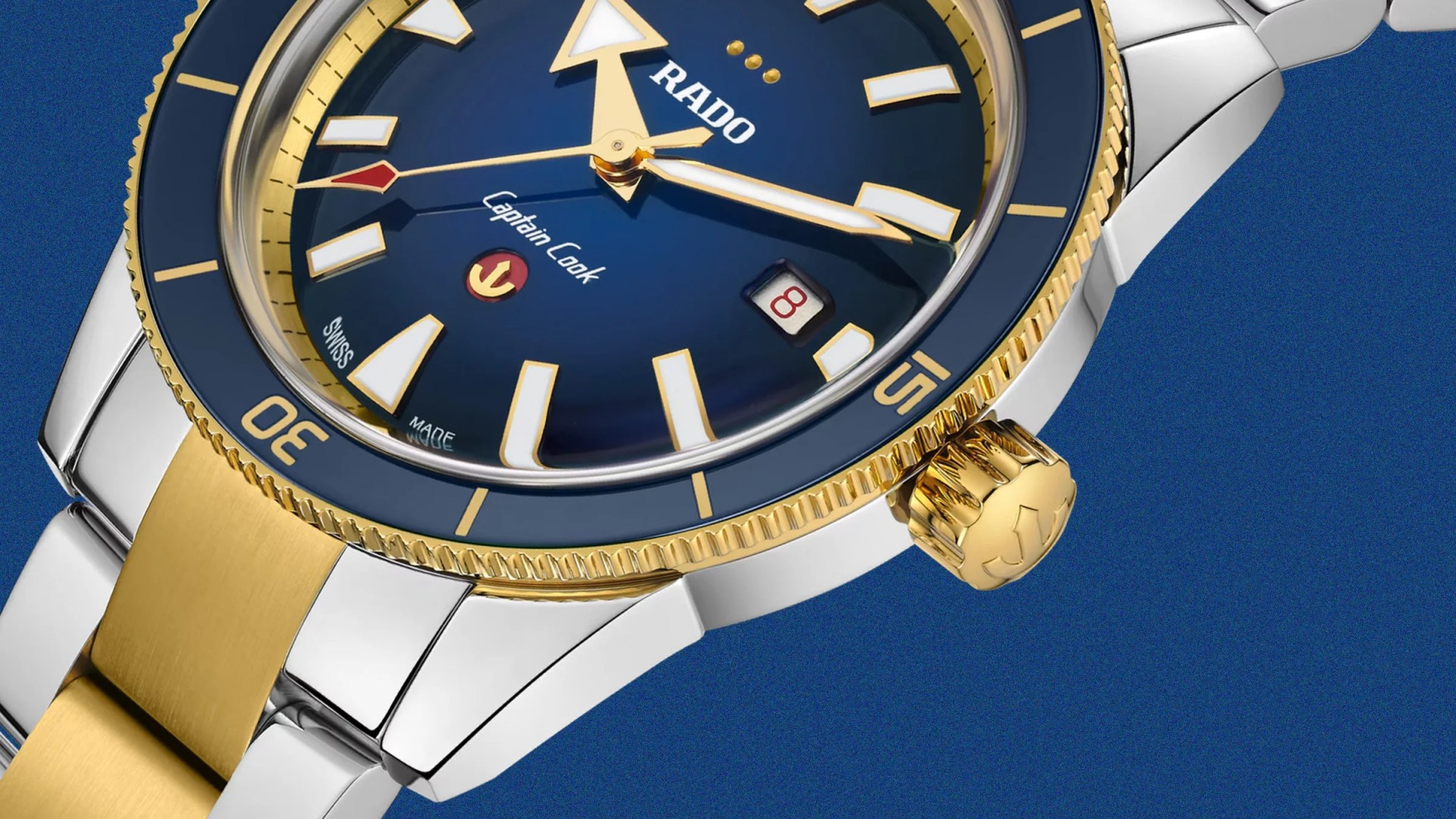 Close-up of a Rado Captain Cook watch with a blue dial, gold and silver case, and date display at 3 o'clock.