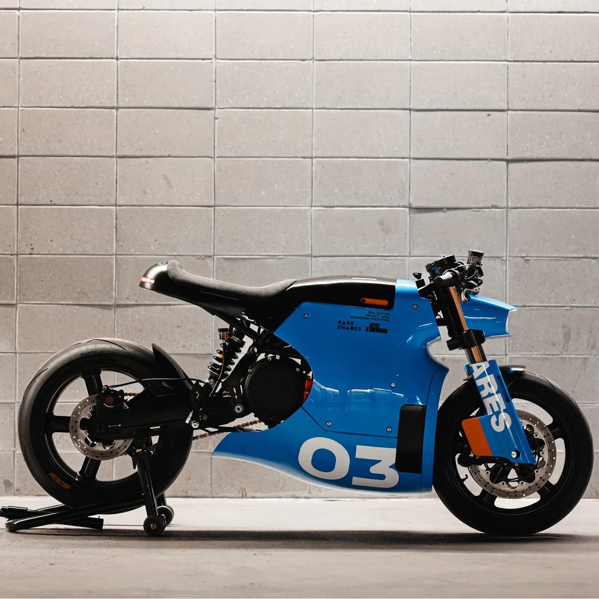 Blue and black electric motorcycle with "ARES" and "03" decals against a gray tiled wall.
