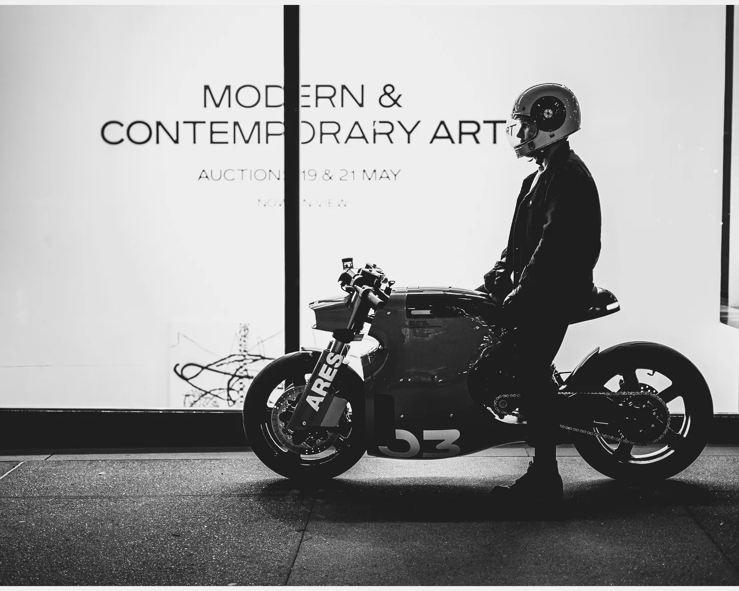 Person wearing a helmet sitting on a motorcycle with "ARES" and "53" on it, in front of a "Modern & Contemporary Art" auction sign.