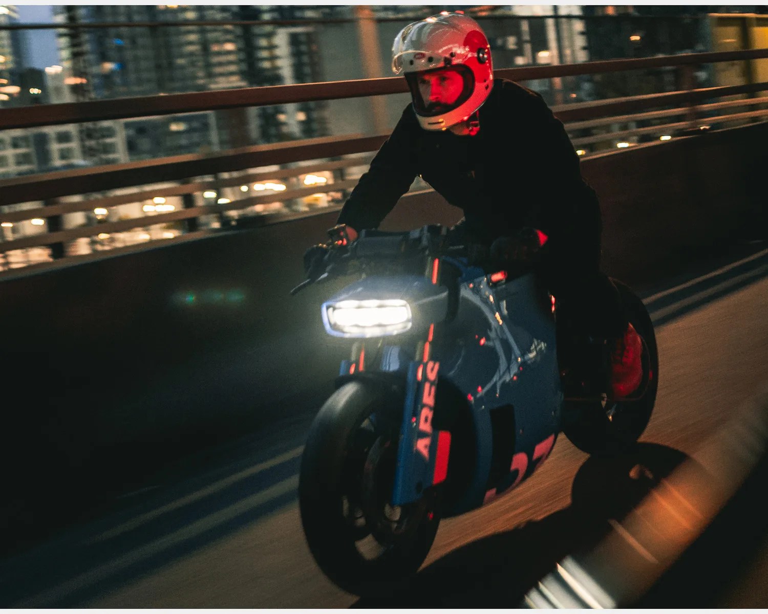 Person wearing a white helmet riding a blue electric motorcycle with bright headlight on a city road at night.