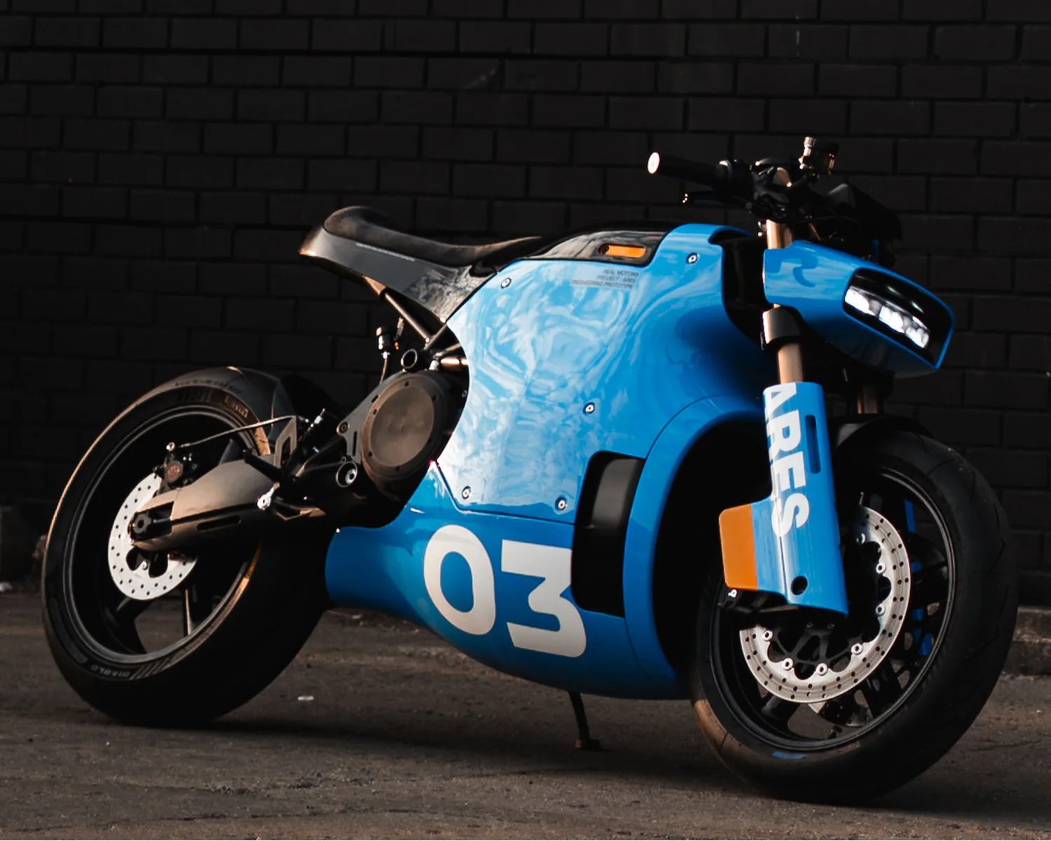 Blue racing motorcycle with number 03 and "ARES" text on front fork, parked on asphalt against black brick wall.