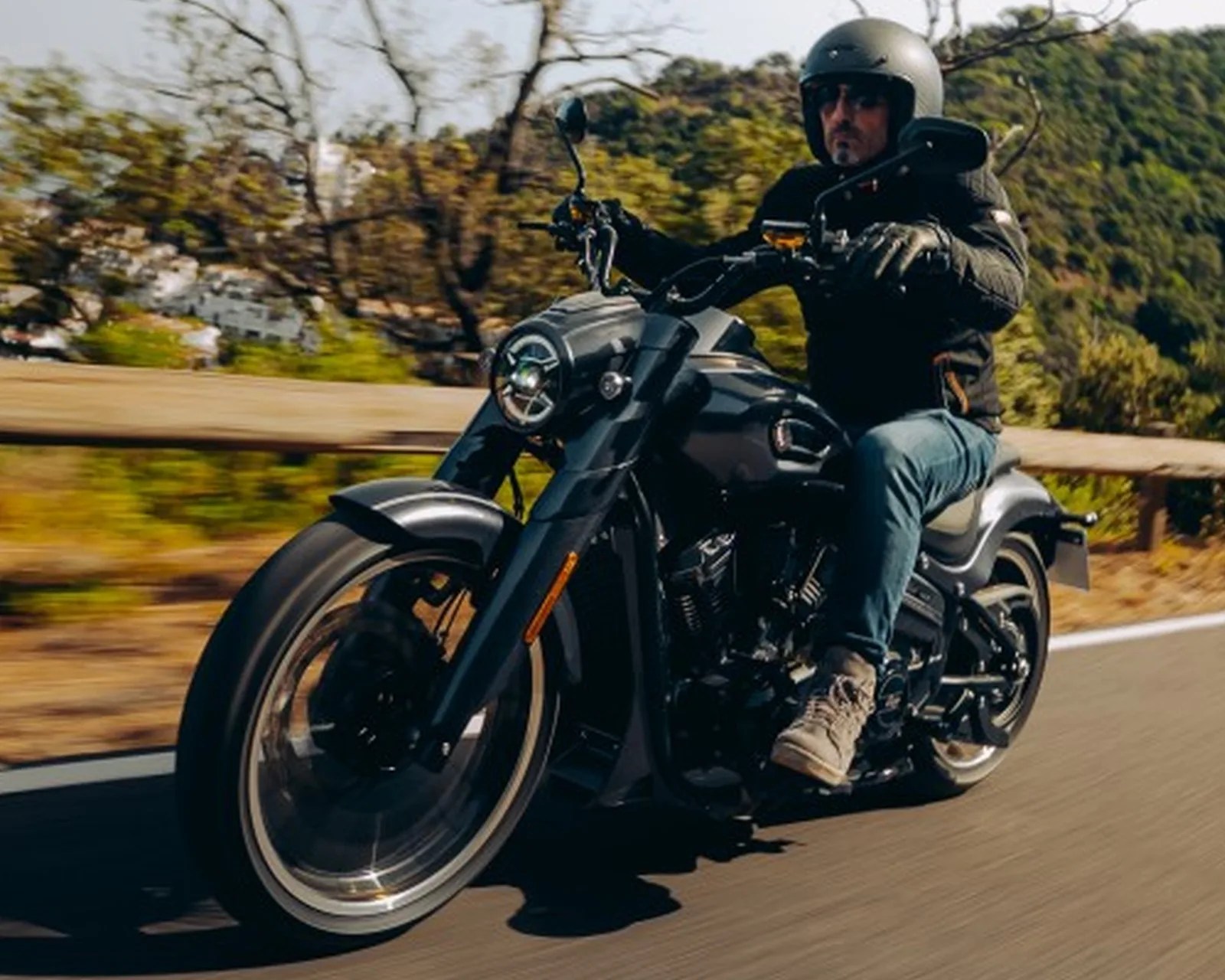 Man wearing a black helmet and jacket riding a black motorcycle on a road with greenery in the background.