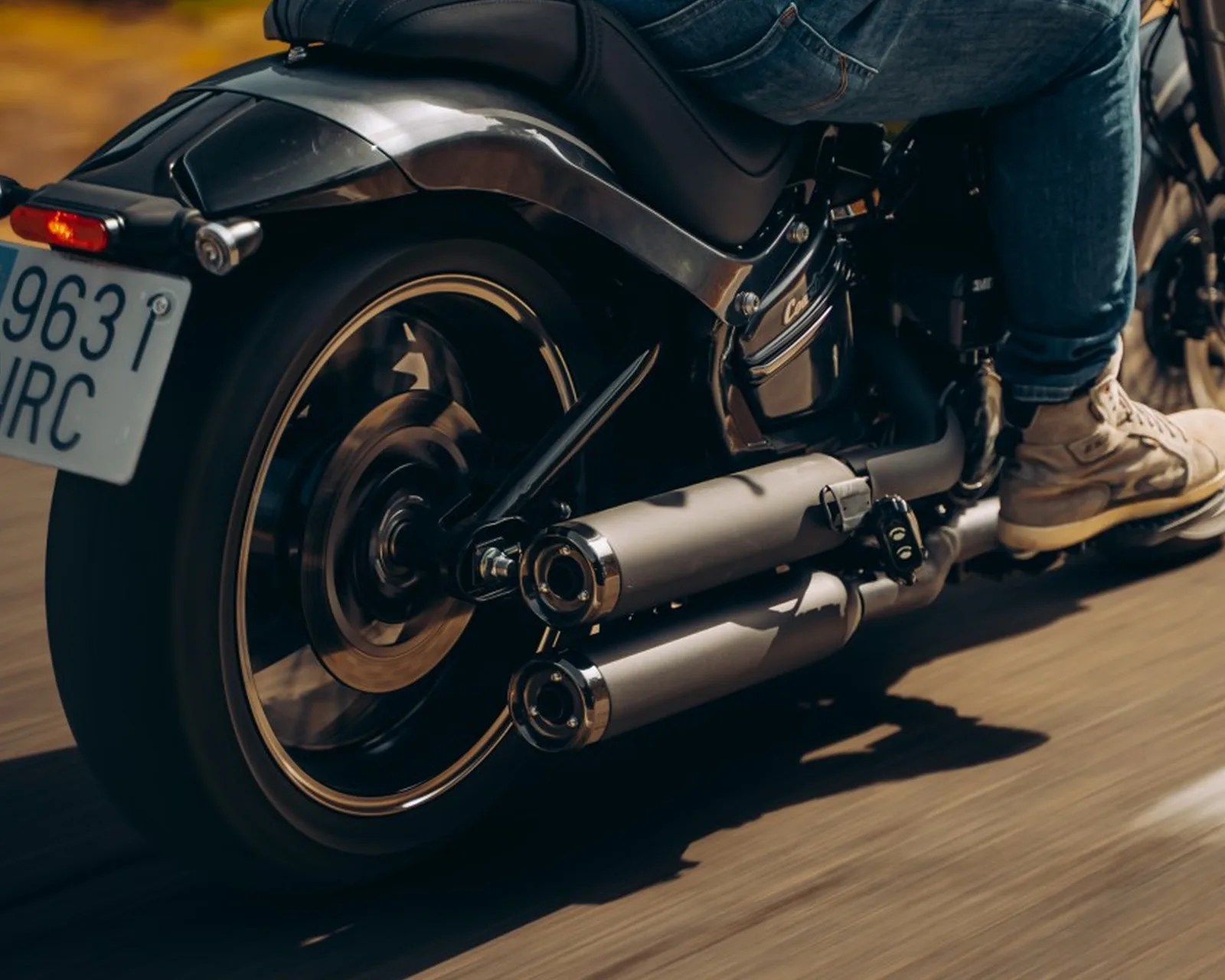 Close-up of a black motorcycle's rear wheel, dual exhaust pipes, and a person wearing beige sneakers and blue jeans riding it.