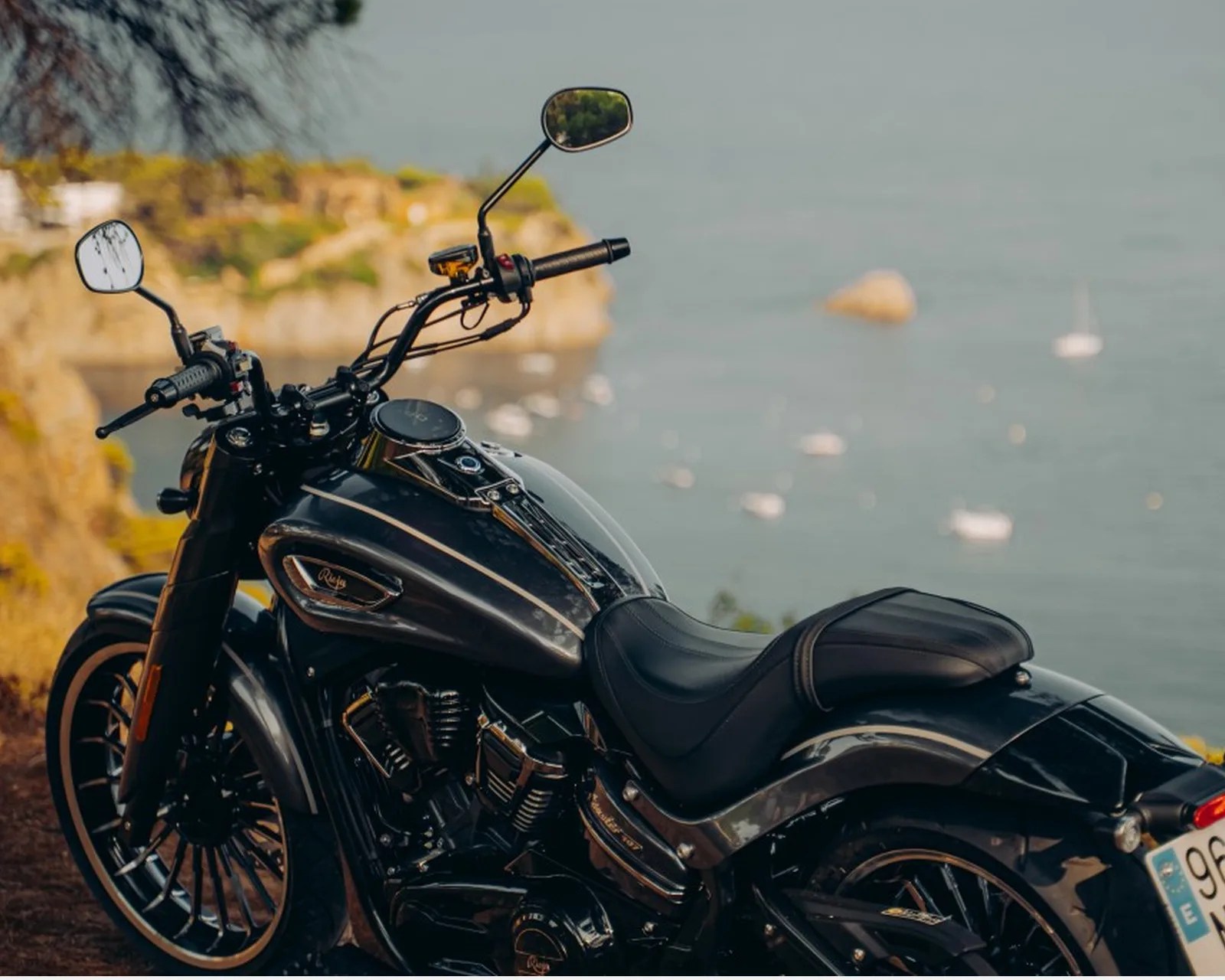 Black cruiser motorcycle parked near a coastal cliff with water and boats in the background.