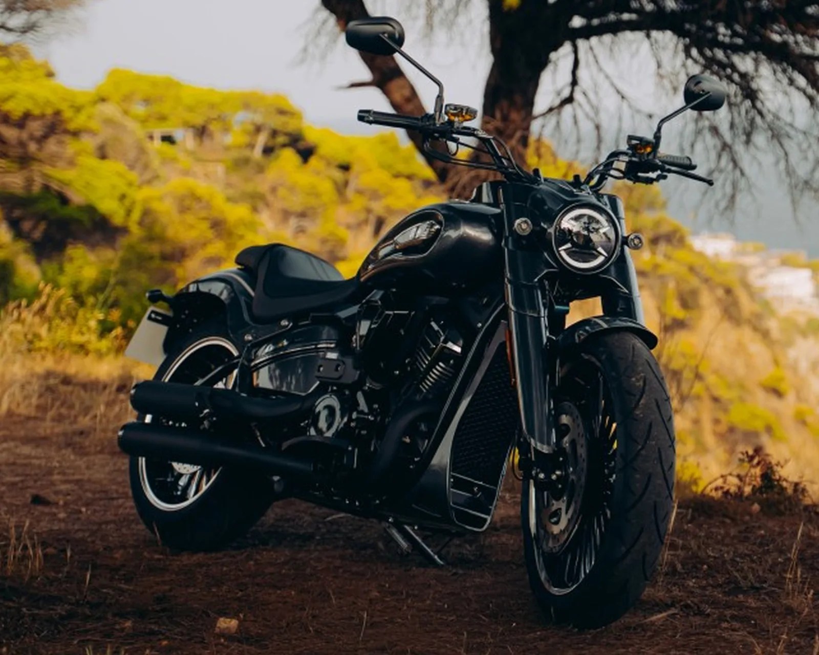 Black cruiser motorcycle with wide tires parked on a dirt path with trees and yellow foliage in the background.