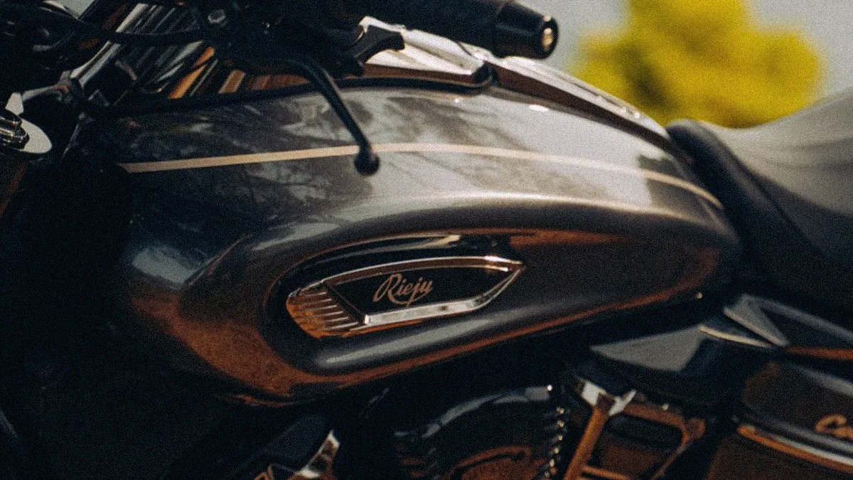Close-up of a dark metallic motorcycle fuel tank with the "Rieju" logo and a thin light stripe on top.
