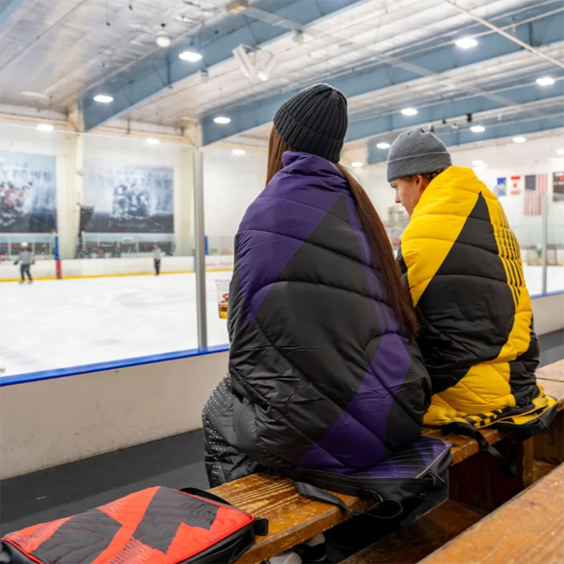 Two people wearing puffer jackets and knit hats sitting on a wooden bench inside an ice rink.