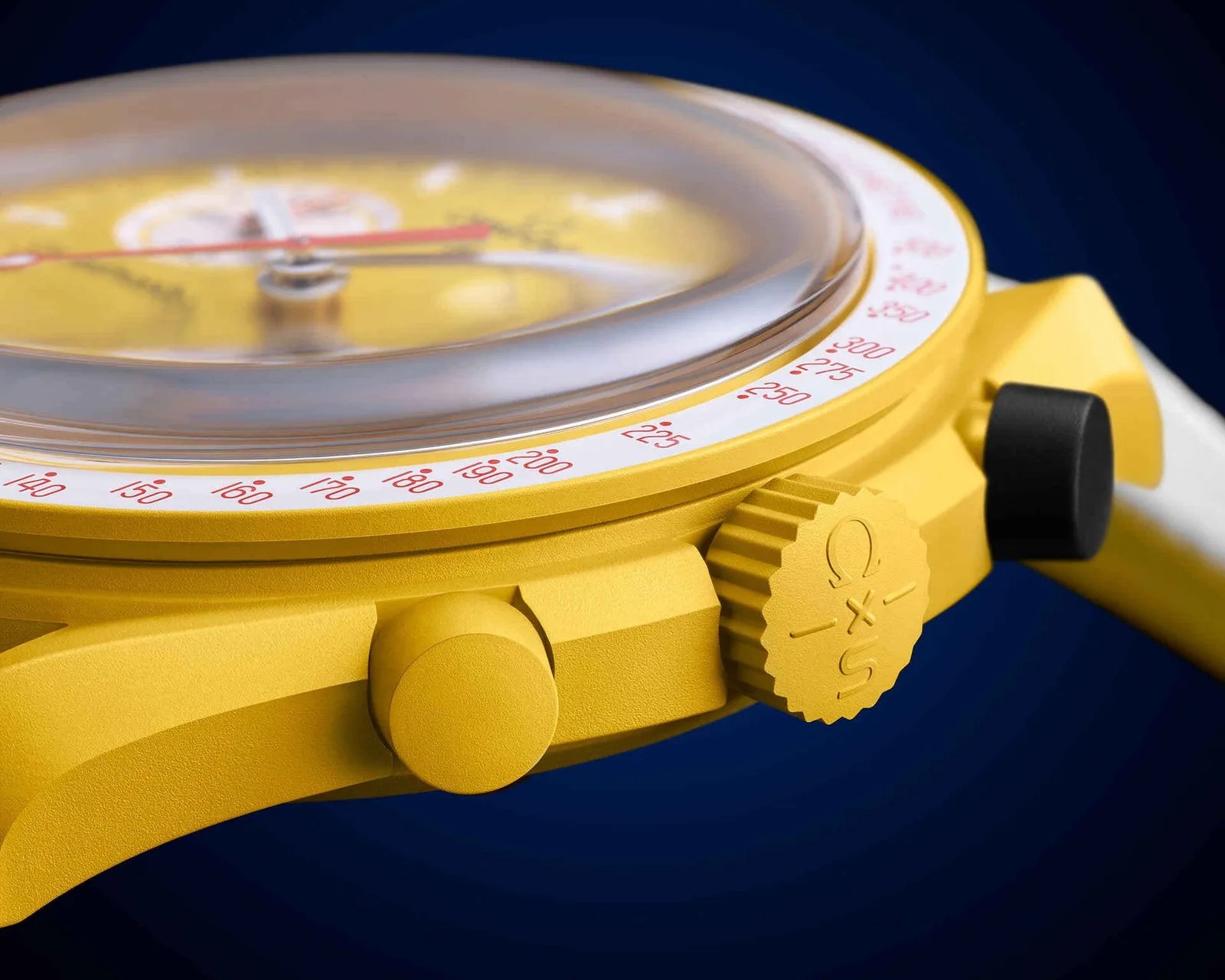 Close-up of a yellow watch case with a textured crown engraved with "Ω x S" and a black pusher against a dark blue background.