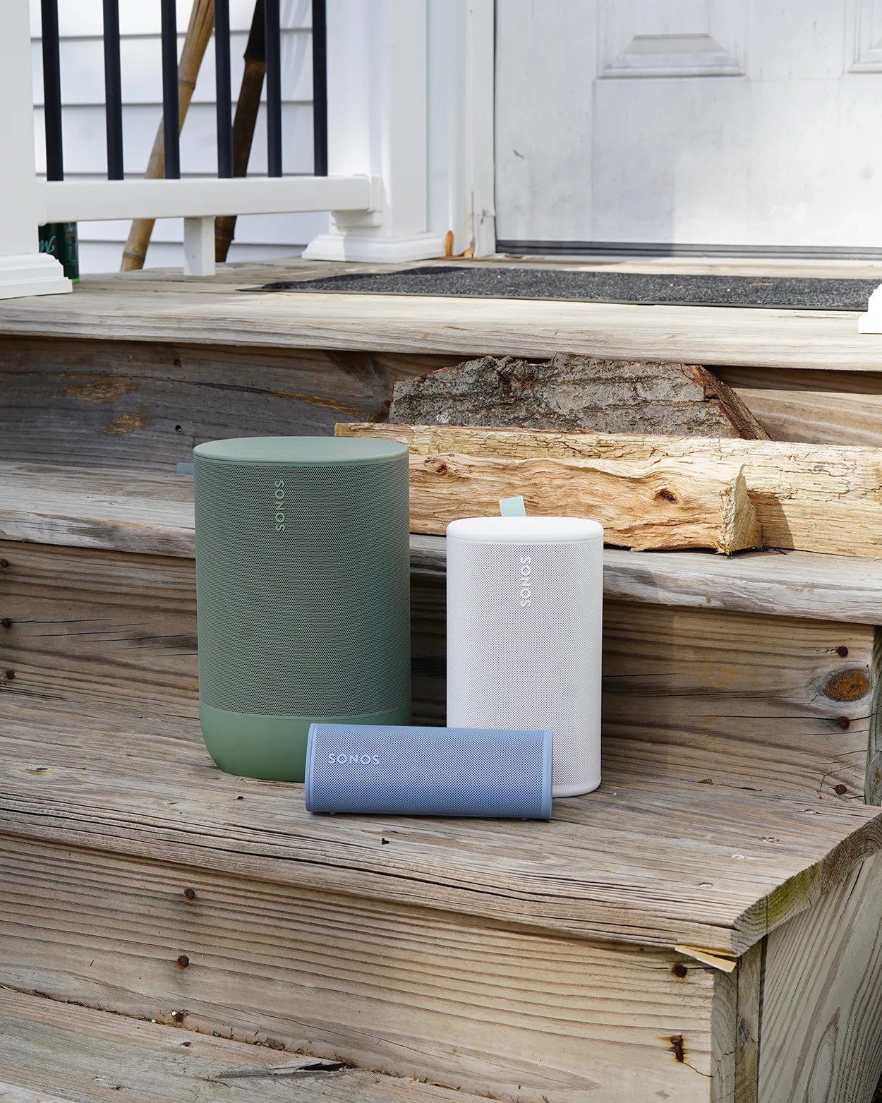 Three Sonos speakers in green, white, and blue on wooden outdoor steps near a white door.