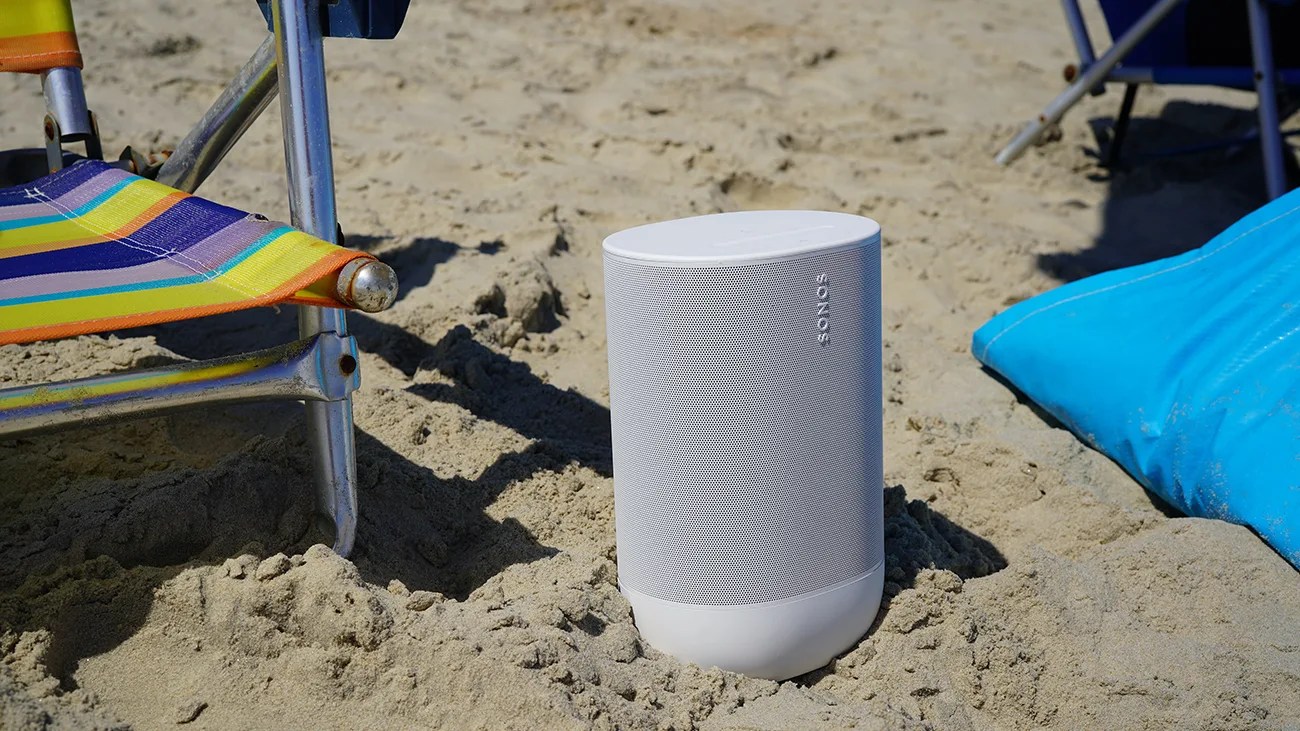 White Sonos speaker standing upright on sandy beach between a colorful striped chair and a blue cushion.