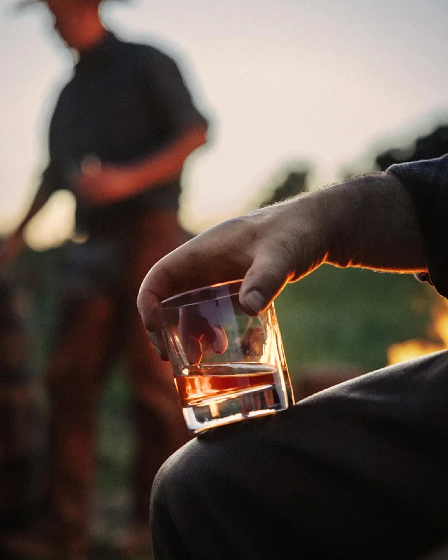 Hand holding a glass with amber liquid, with a blurred person in the background at sunset.