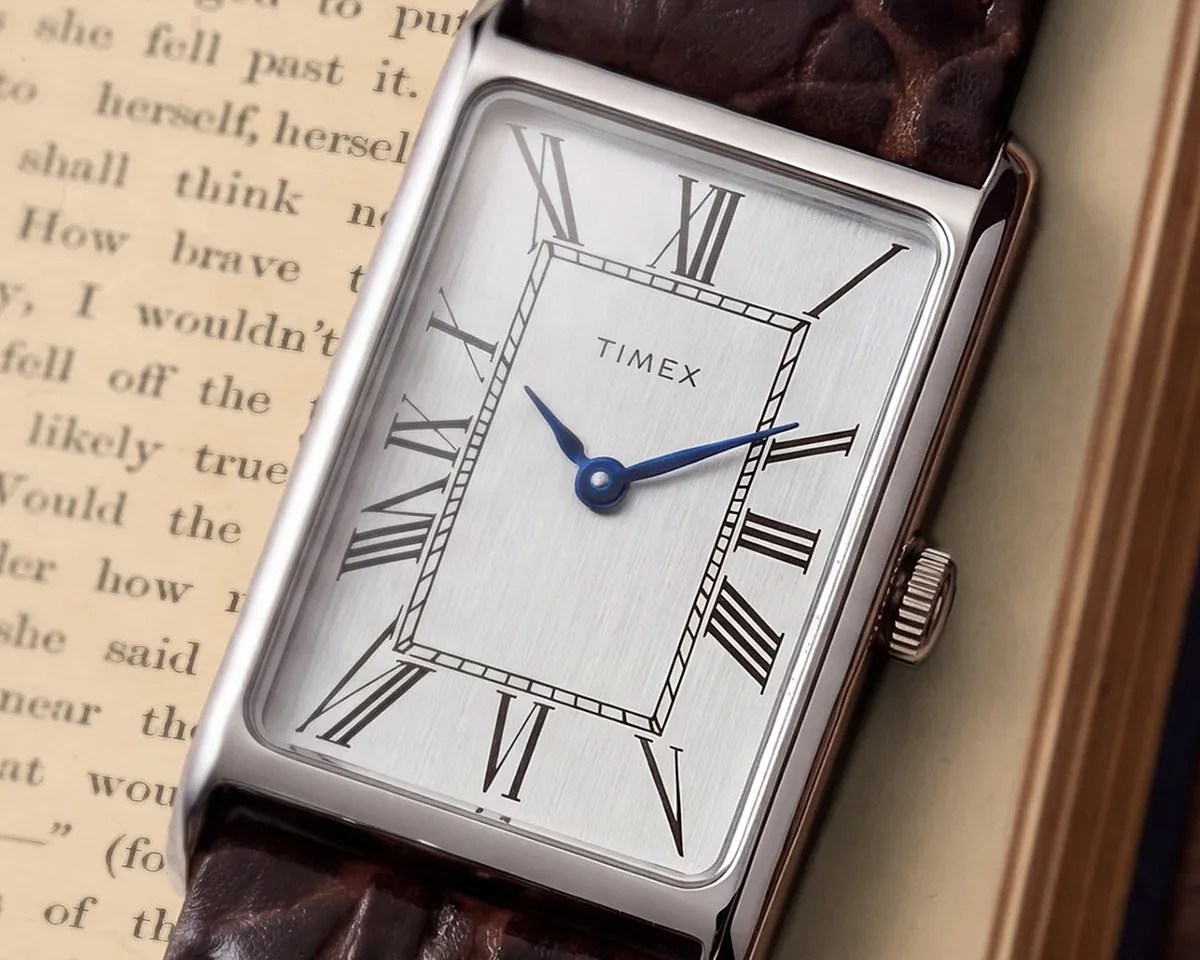 Rectangular Timex watch with silver dial, black Roman numerals, blue hands, and brown leather strap on an open book.