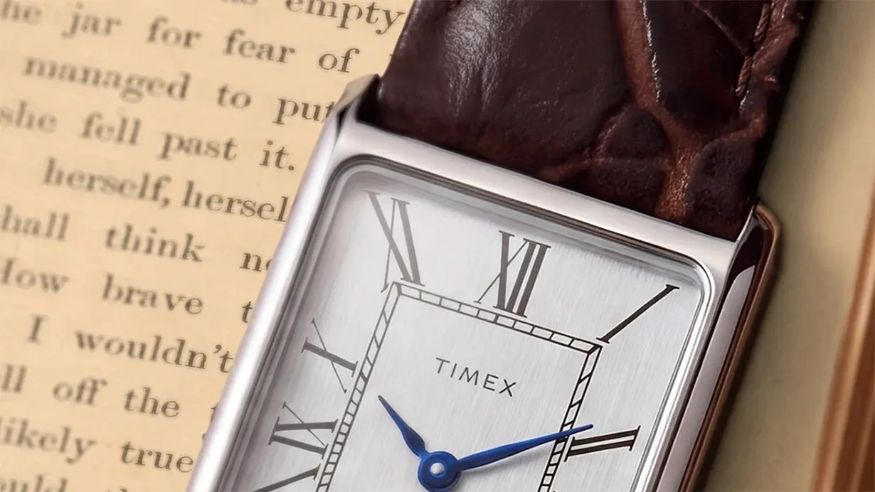 Close-up of a Timex watch with a rectangular silver face, blue hands, Roman numerals, and a brown leather strap.