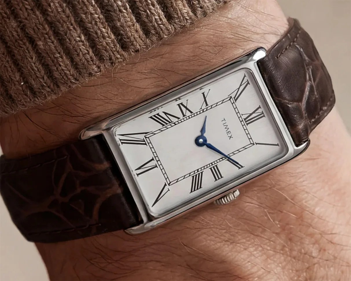 Rectangular silver Timex wristwatch with white dial, black Roman numerals, blue hands, and brown leather strap worn on a wrist.