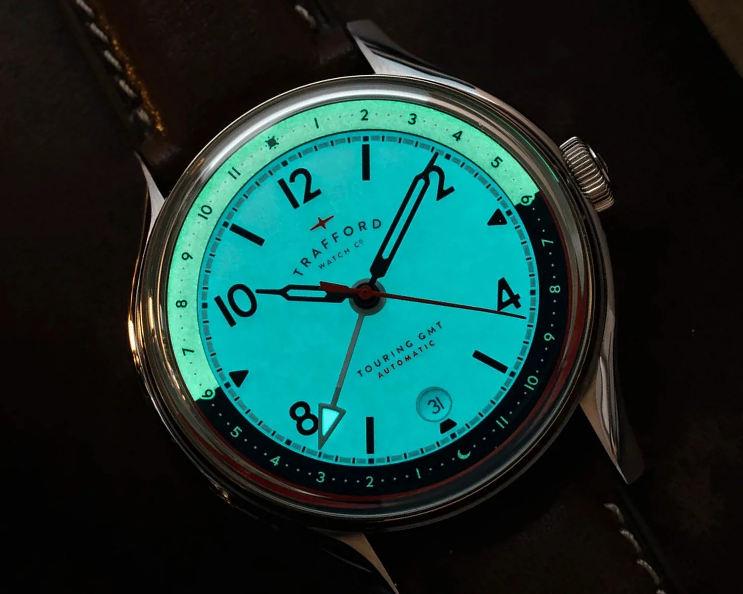 Close-up of a glowing turquoise watch dial with black numerals, silver case, and brown leather strap.