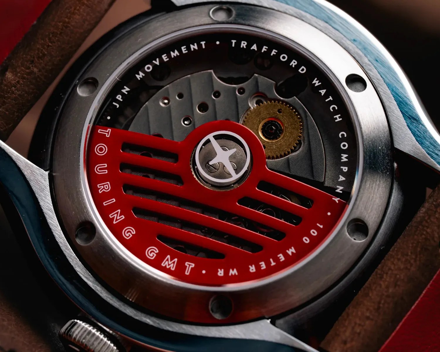 Close-up of a watch movement with a red rotor labeled "Touring GMT" inside a stainless steel case.