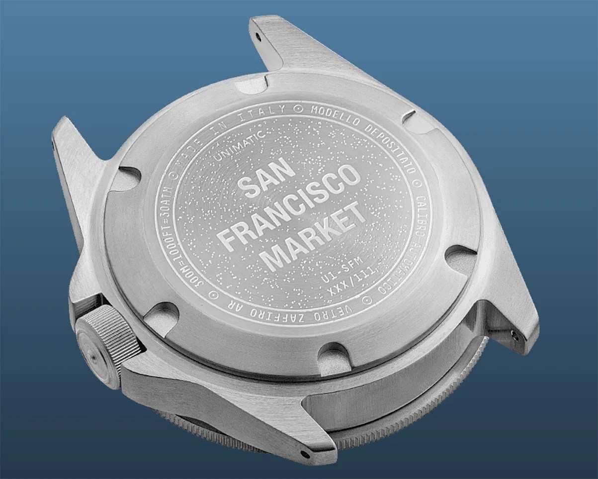 Brushed stainless steel watch case back engraved with "San Francisco Market" and other text on a blue background.