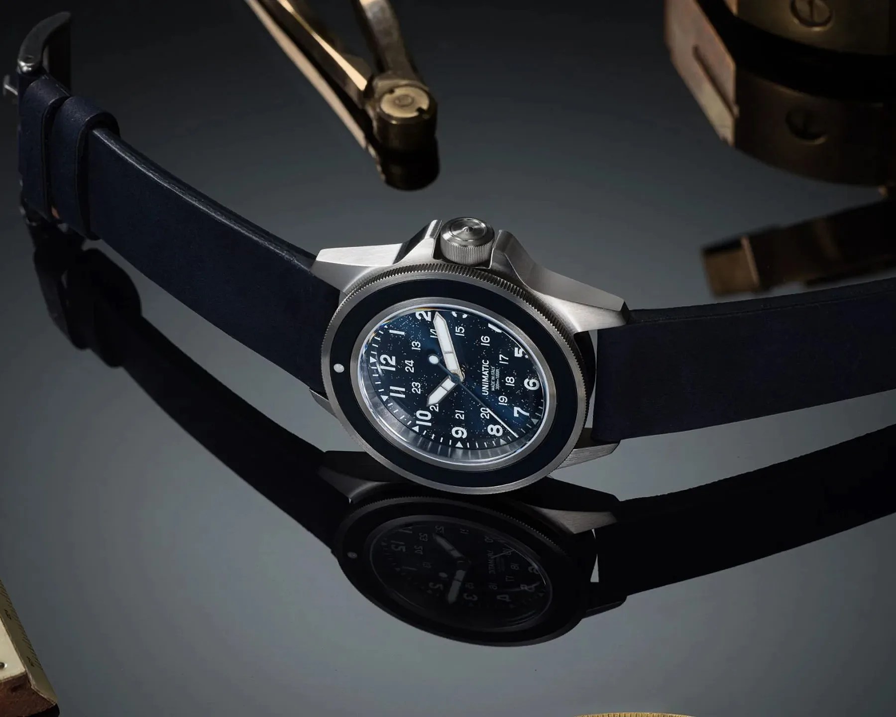Stainless steel wristwatch with a black bezel, navy blue dial, white numerals, and navy blue strap on a reflective surface.