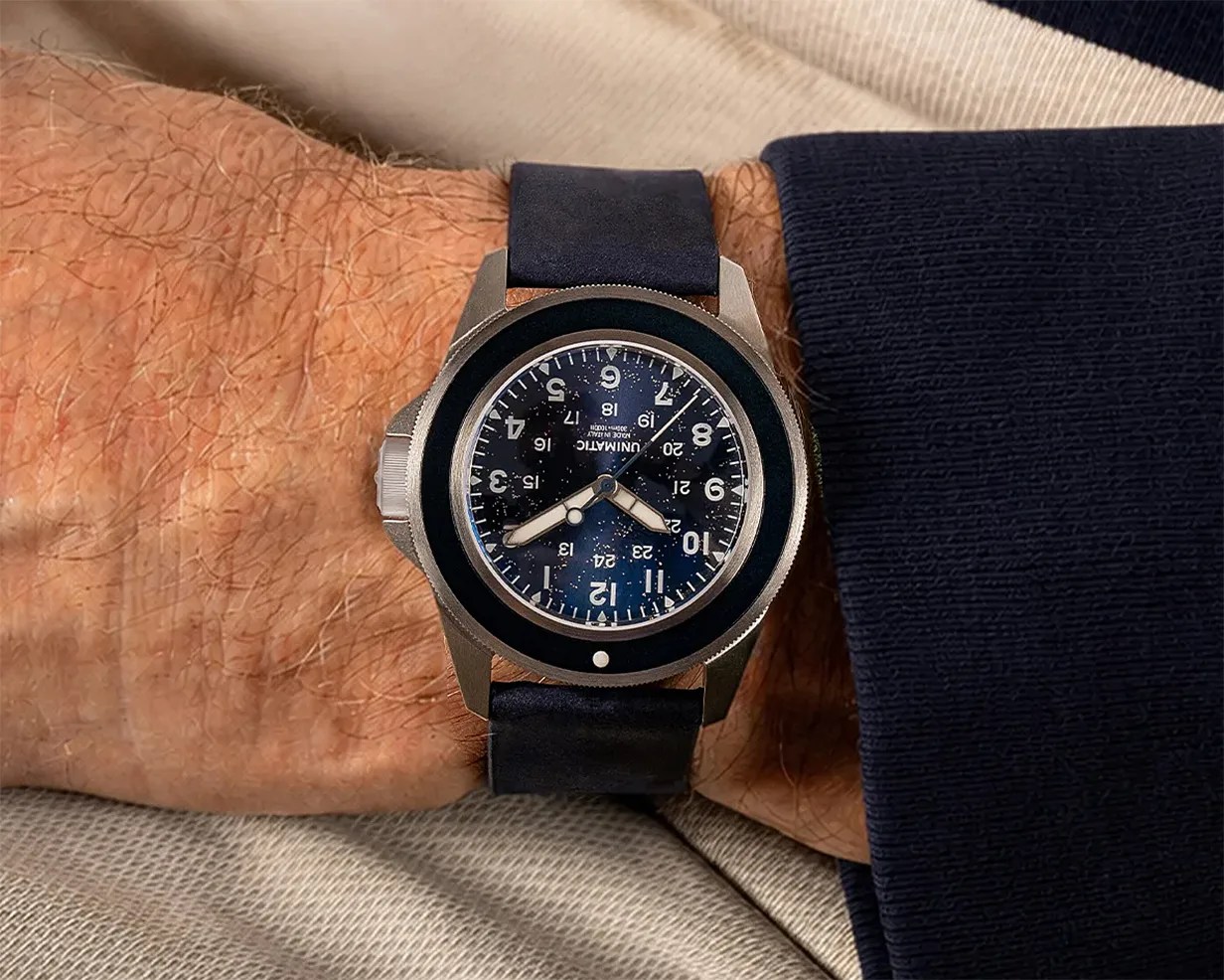 Wristwatch with black dial, white numerals, and black leather strap worn on a wrist with a navy sleeve.