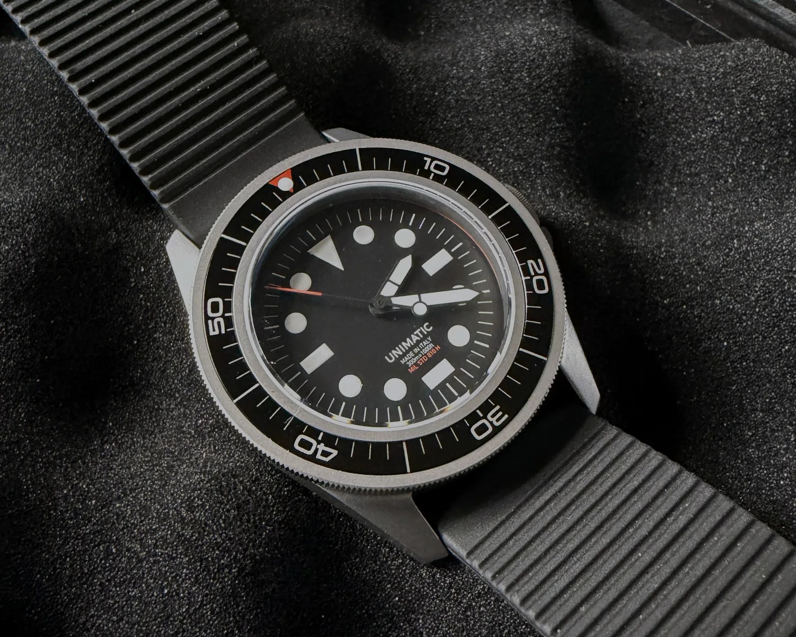 Black Unimatic dive watch with white markers and a black ribbed rubber strap on a dark textured surface.