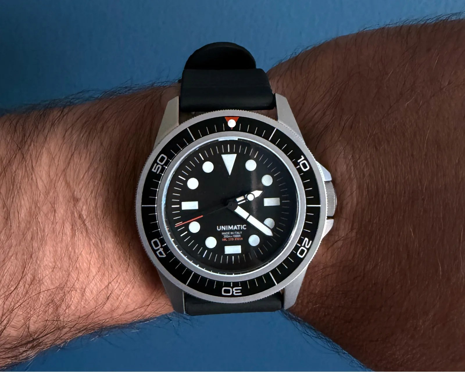 Black Unimatic wristwatch with white markers and a black bezel on a wrist against a blue background.