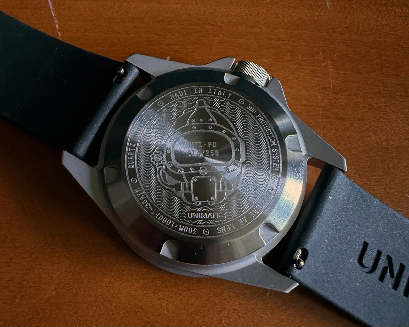 Back of a stainless steel Unimatic watch with engraved diver helmet design and black strap on a wooden surface.