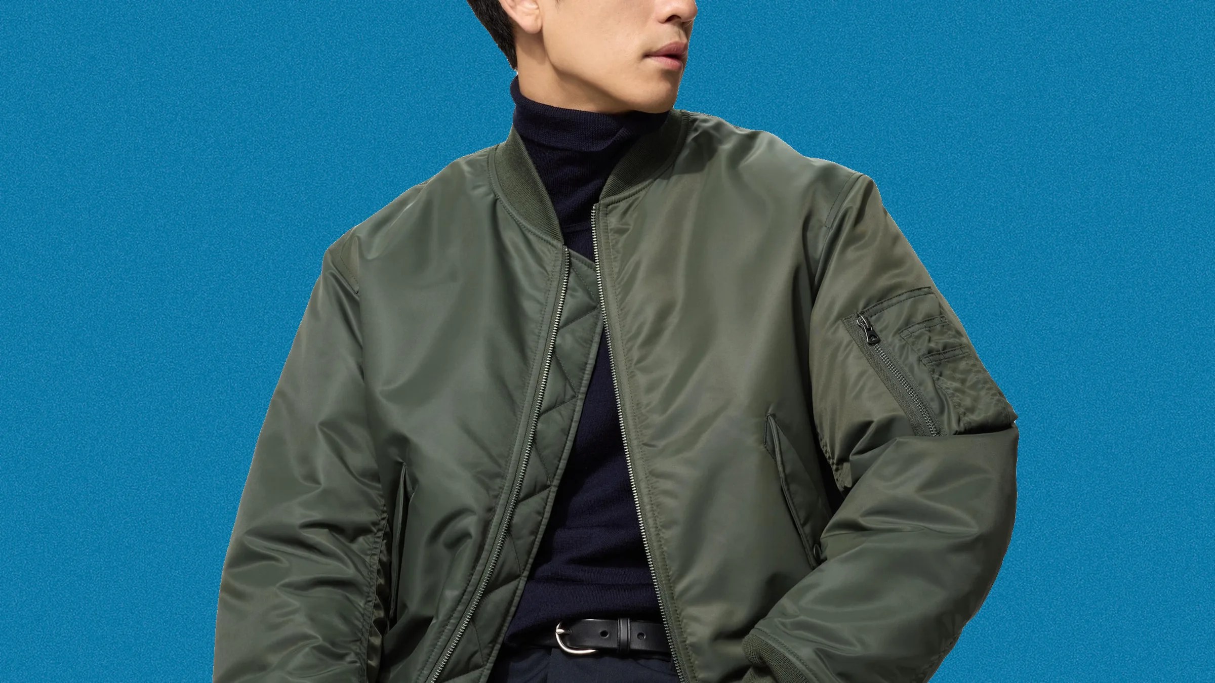 Olive green bomber jacket with zippered arm pocket worn over a black turtleneck and black belt.