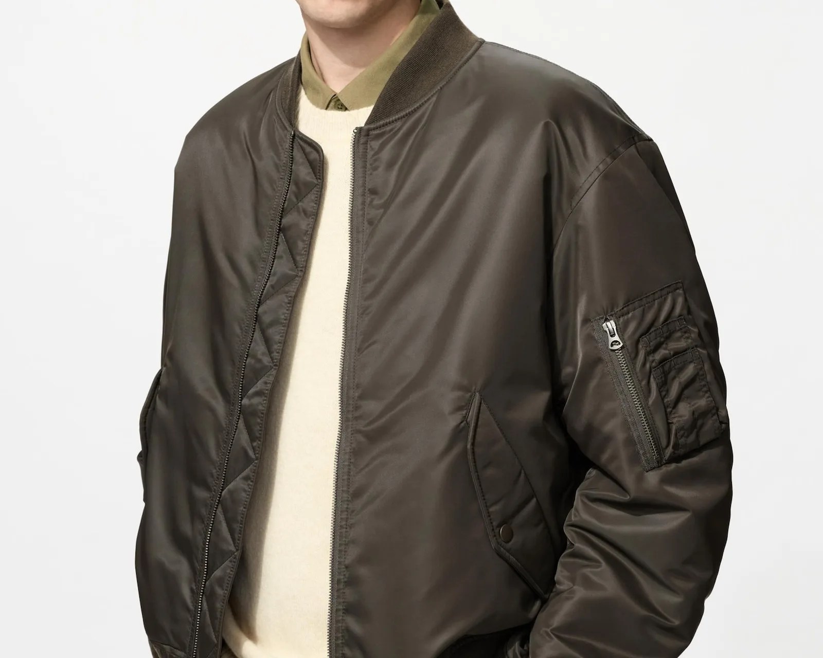 Dark olive green bomber jacket with zippered pocket on sleeve worn over a cream sweater and olive shirt.