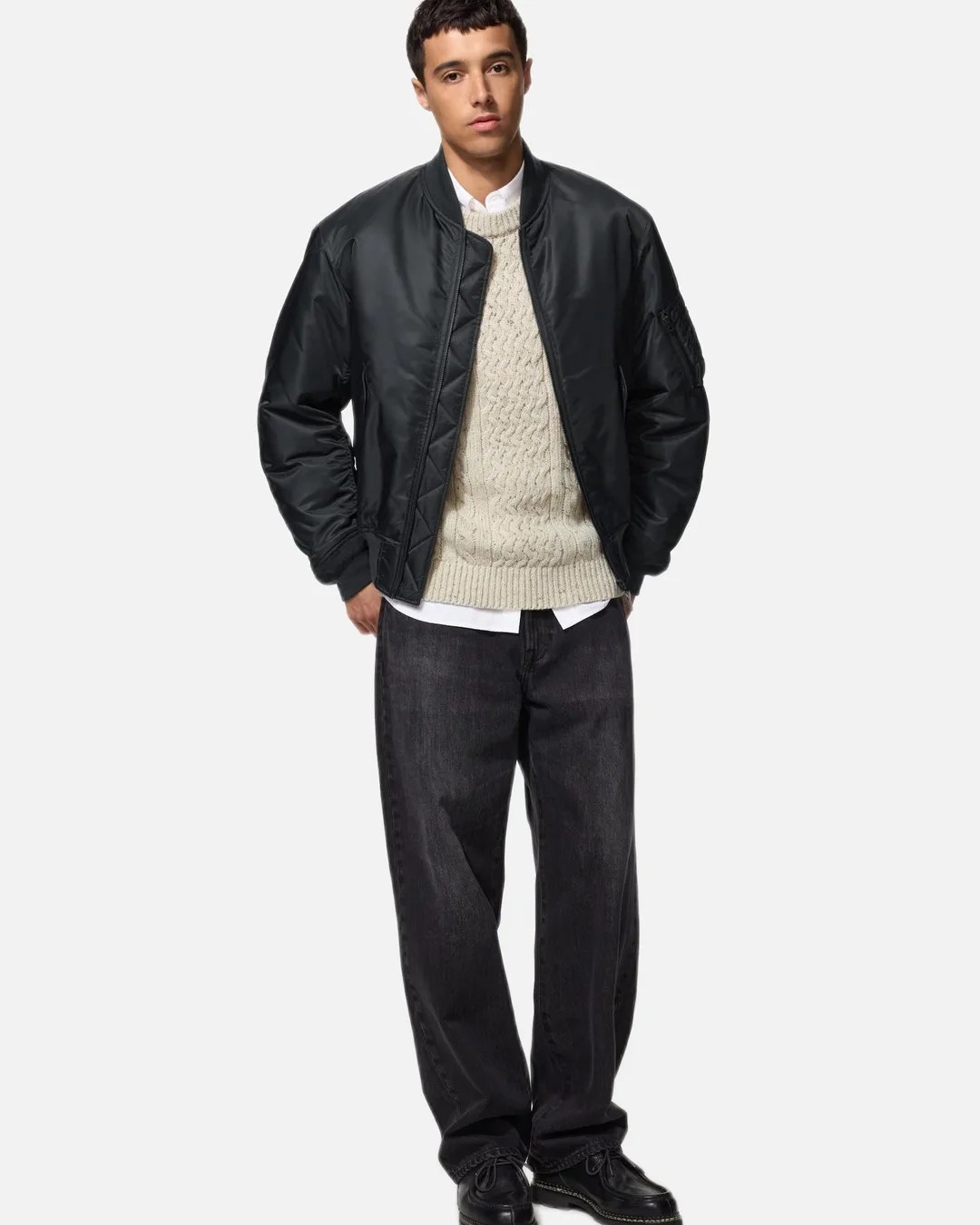 Man wearing a black bomber jacket over a cream cable-knit sweater, white shirt, loose black jeans, and black shoes.