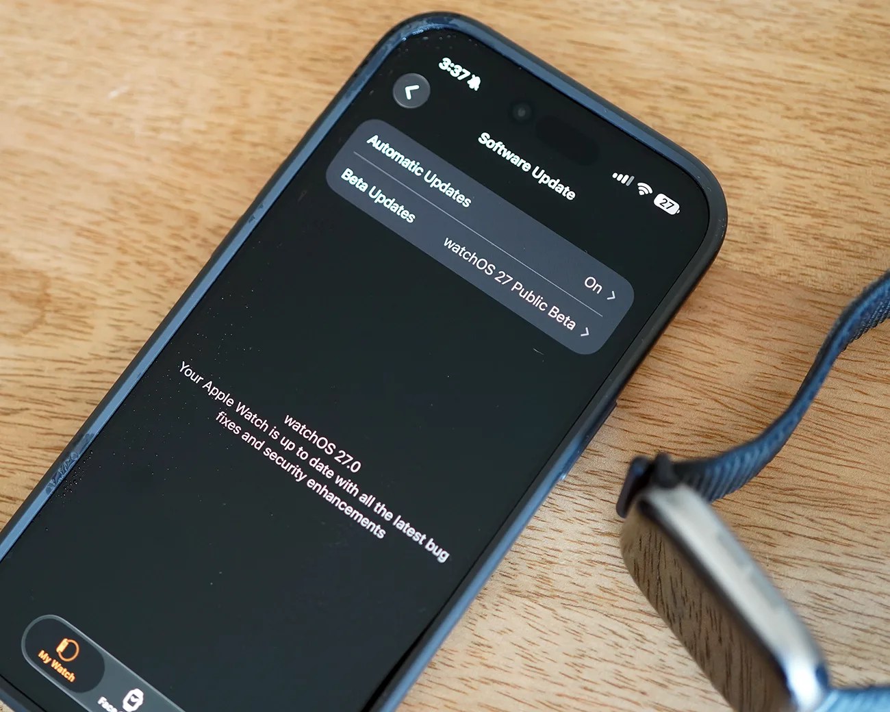 Smartphone displaying watchOS 27.0 software update screen next to an Apple Watch with a blue woven band on a wooden surface.