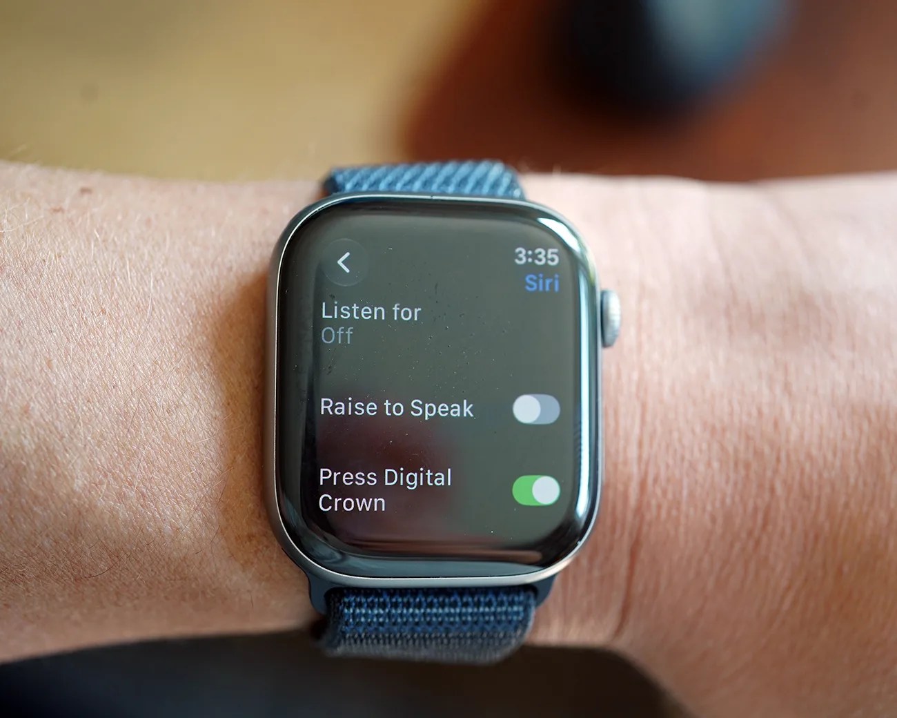 Smartwatch with blue fabric band showing Siri settings screen with "Raise to Speak" off and "Press Digital Crown" on.