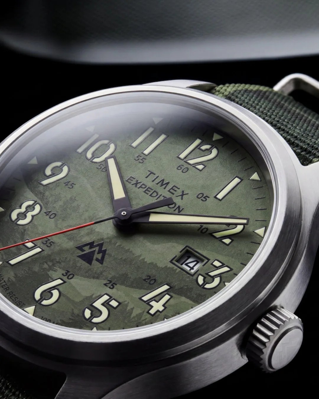 Close-up of a Timex Expedition watch with a green camo dial, luminous hands, date window, red second hand, and green fabric strap.