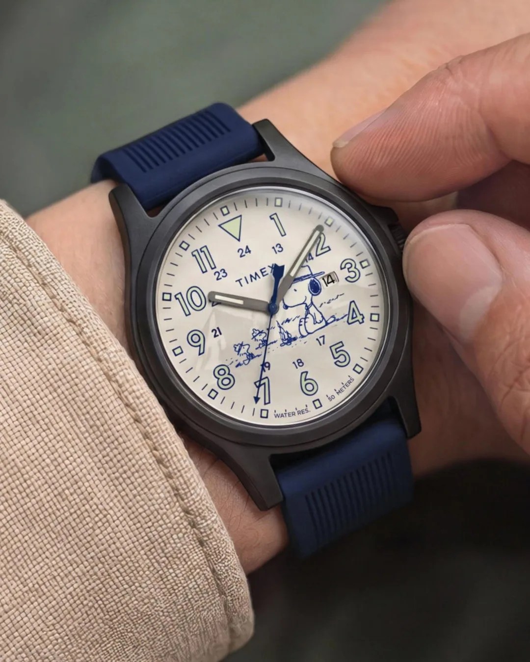 Black Timex wristwatch with blue numerals and Snoopy illustration on white dial, worn with a blue strap.