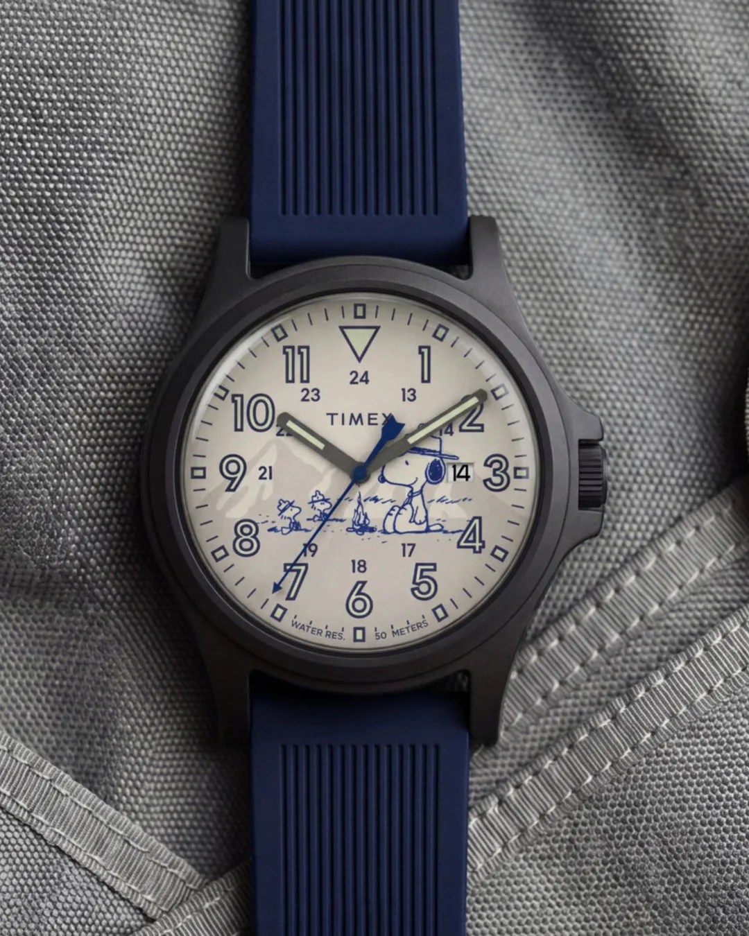Timex wristwatch with black case, white dial featuring Snoopy illustration, blue numerals, and blue silicone strap on gray fabric.