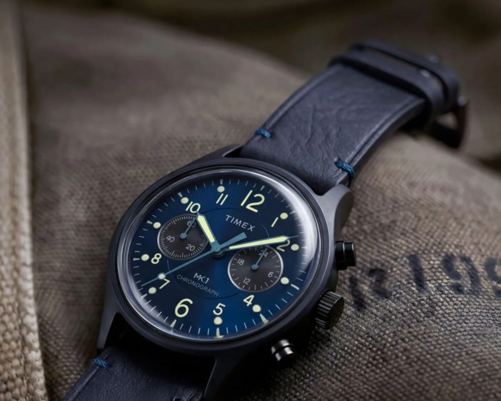 Black Timex MK1 chronograph watch with blue dial and leather strap on textured fabric.