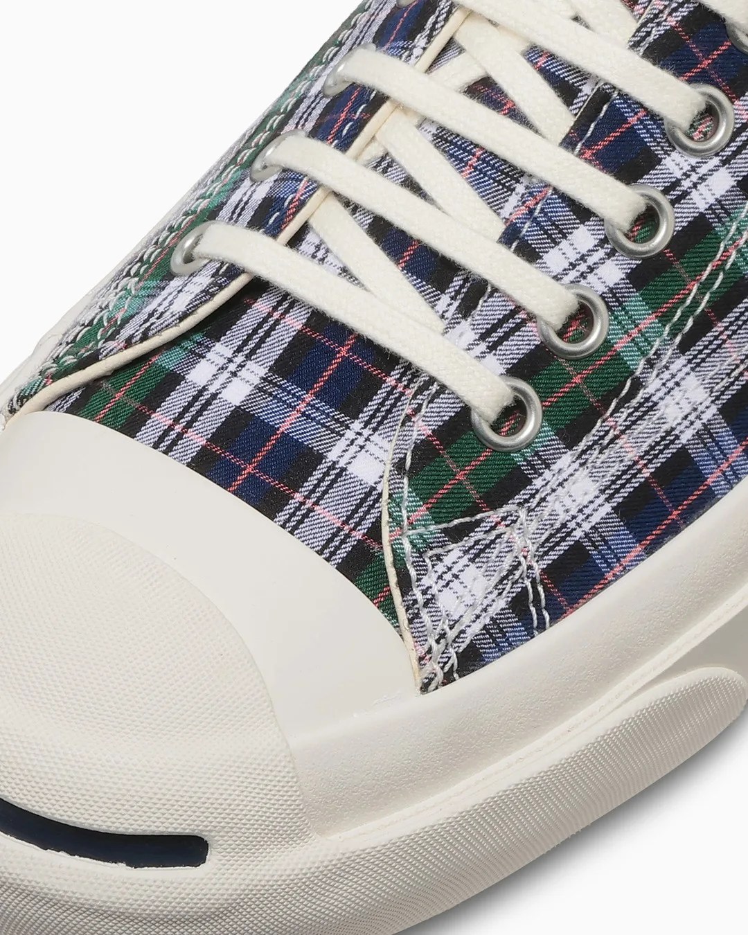 Close-up of a sneaker with white rubber toe cap and sole, featuring green, navy, black, and white plaid fabric upper with white laces.