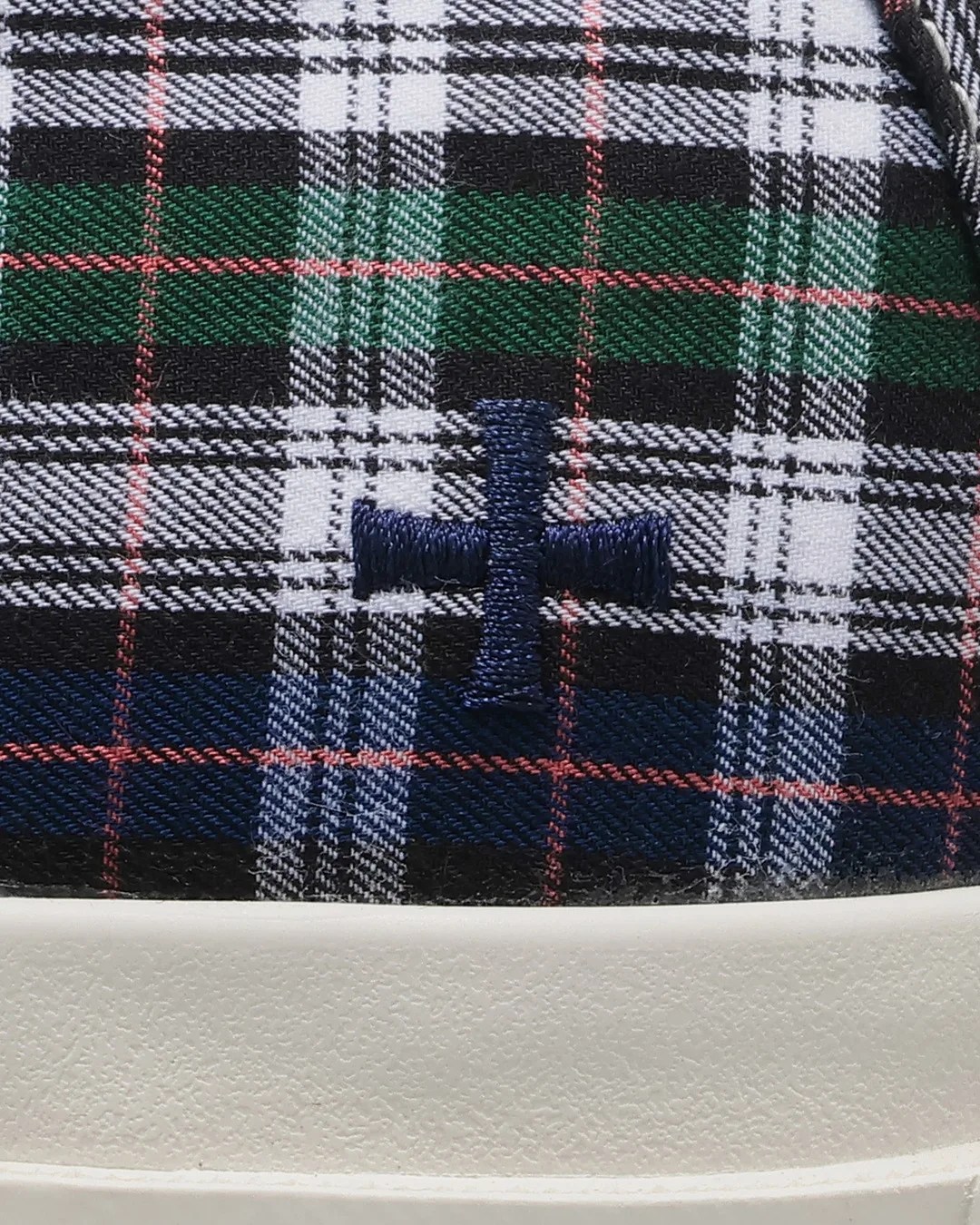 Close-up of a plaid fabric with green, navy, black, white, and red lines, featuring a navy embroidered cross above a white textured sole.