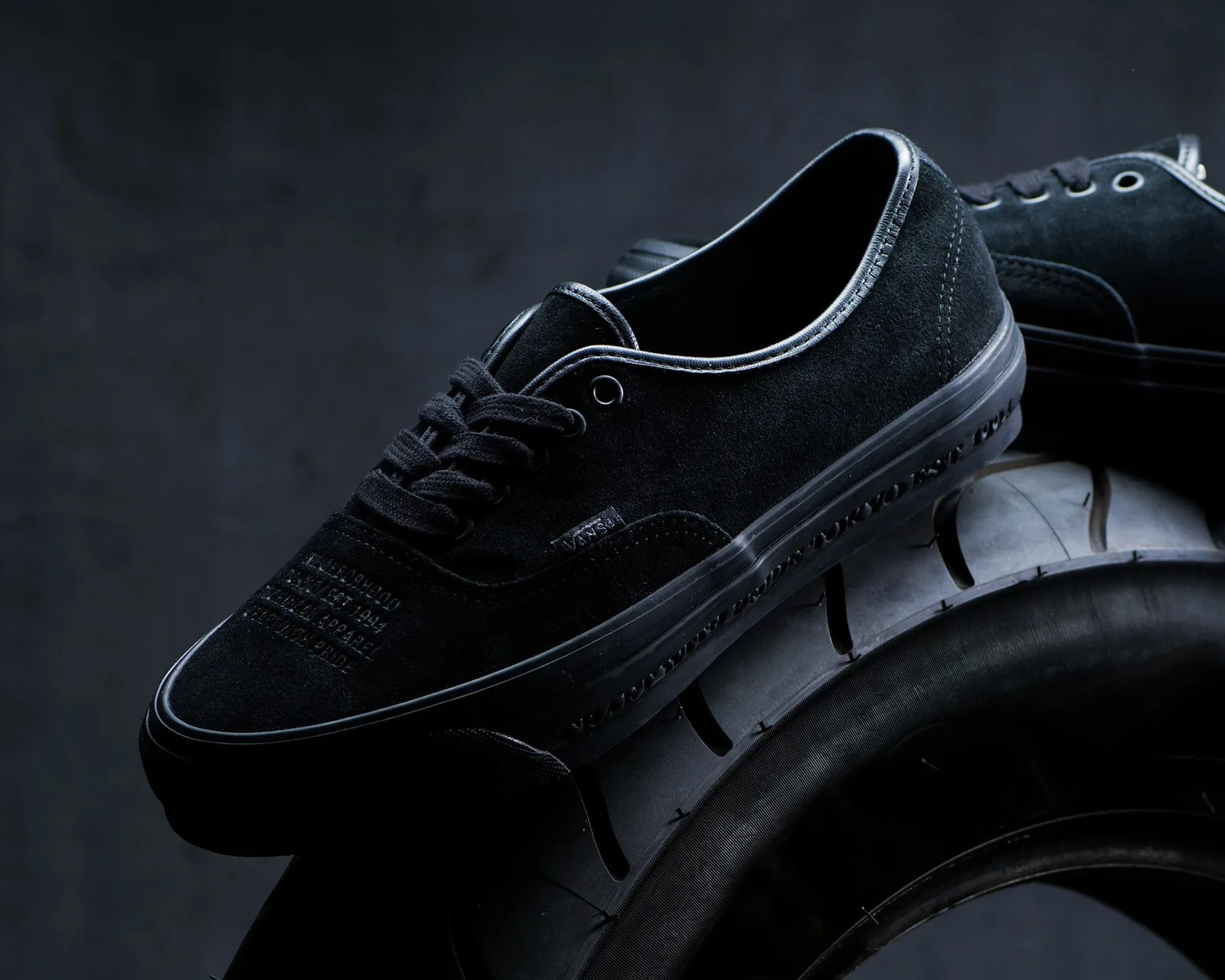 Black Vans suede sneakers with black laces resting on a black tire against a dark background.