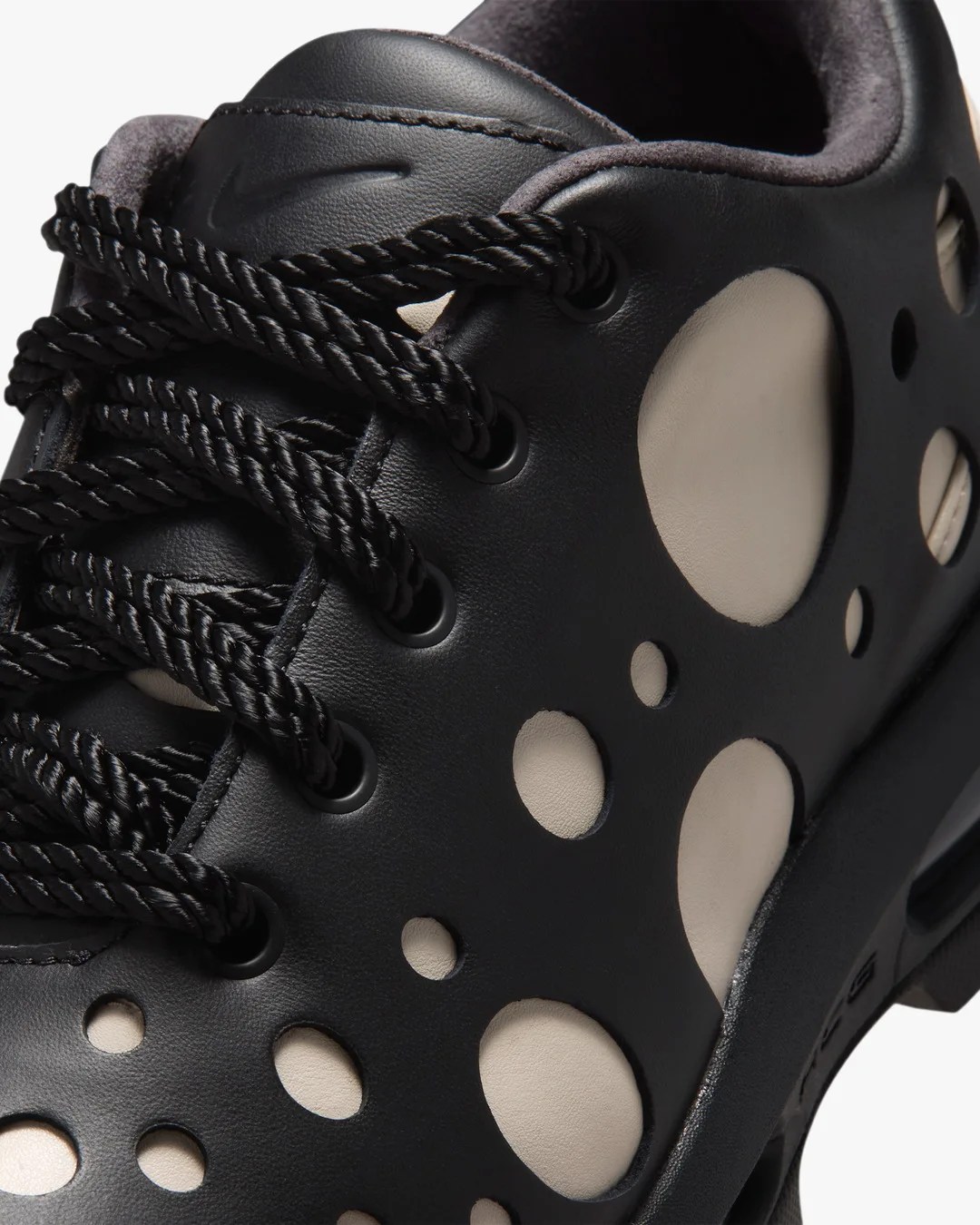 Close-up of a black sneaker with thick black laces and multiple circular cutouts revealing a beige inner layer.