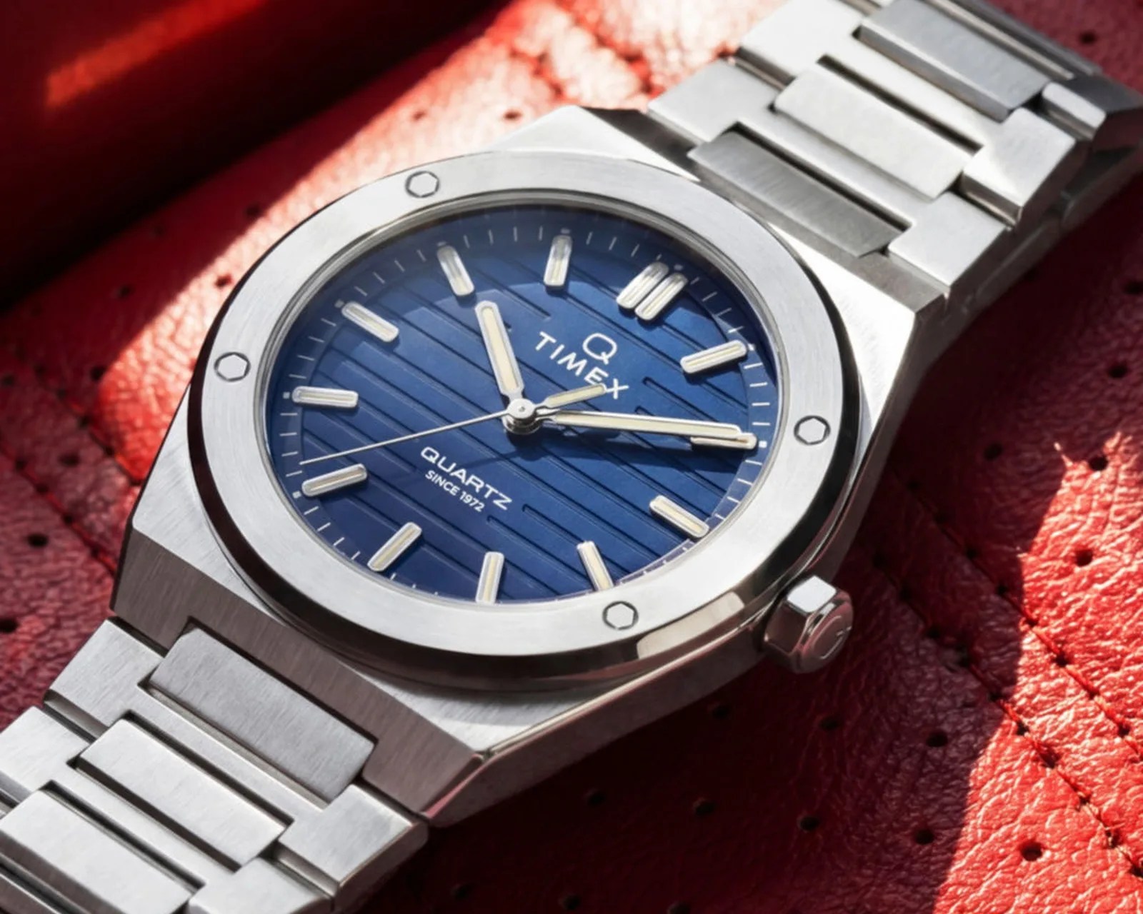 Silver stainless steel wristwatch with a blue textured dial and luminous markers on a red perforated leather surface.