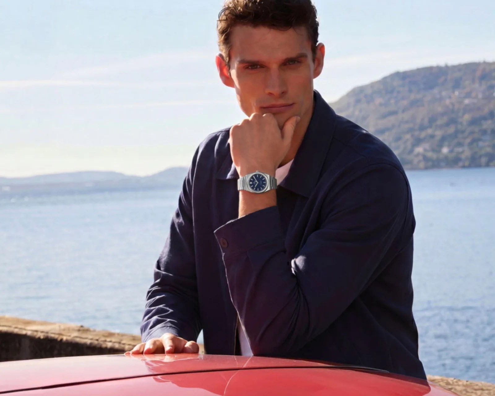 Man wearing a silver wristwatch with a blue dial and metal band, resting his arm on a red surface by the water.