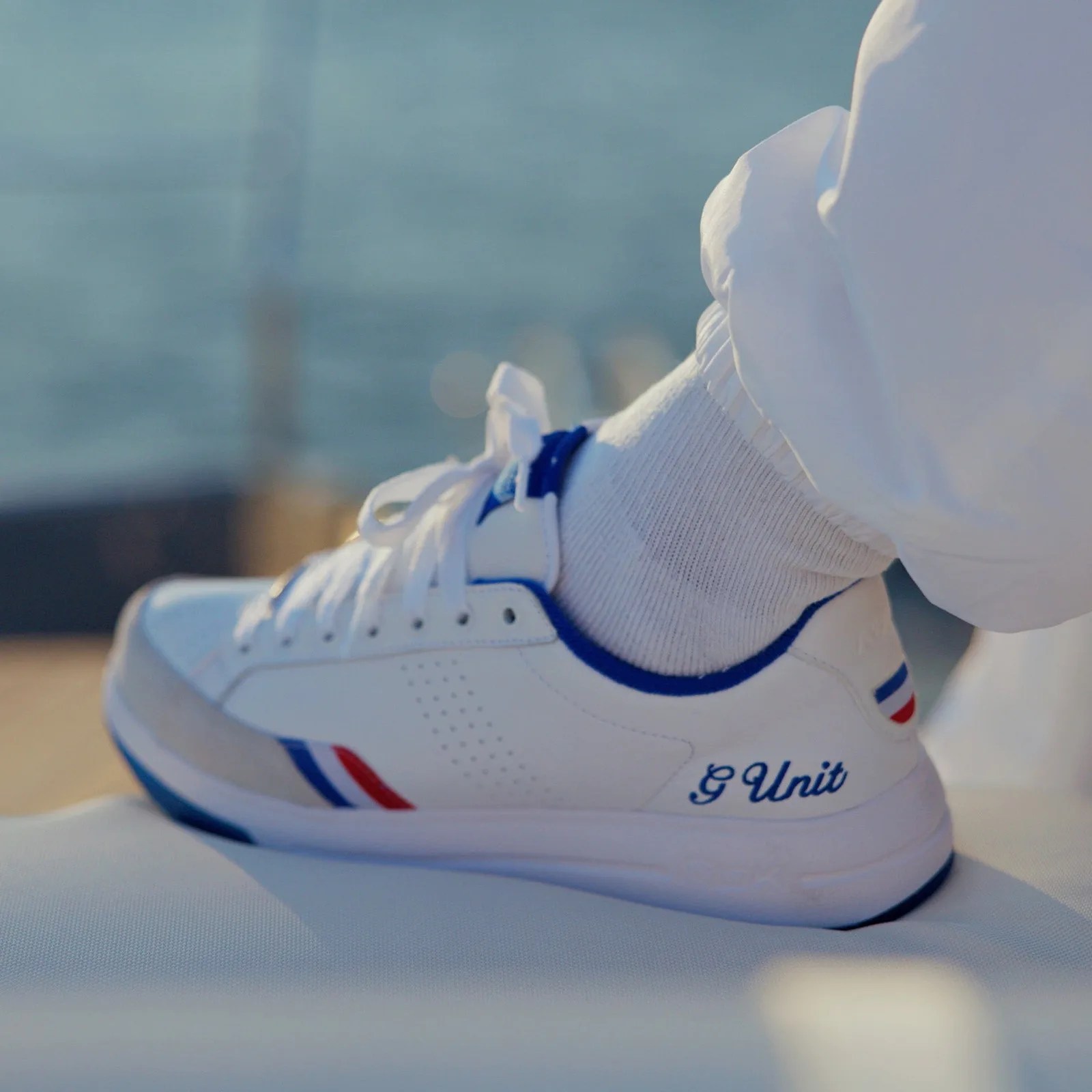 White sneaker with blue and red accents and "G Unit" embroidered on the side, worn with white socks and pants.