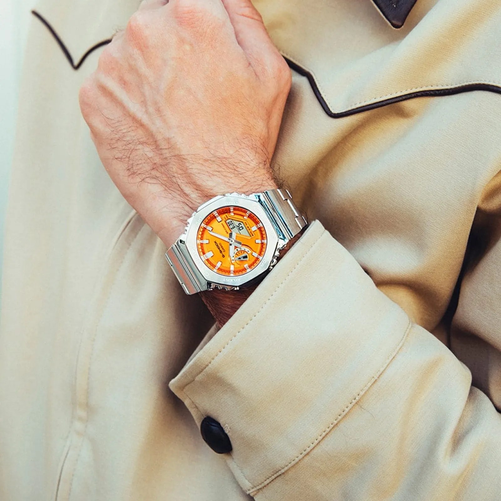 Silver metal wristwatch with an orange dial and digital date display worn on a wrist over a beige jacket sleeve.