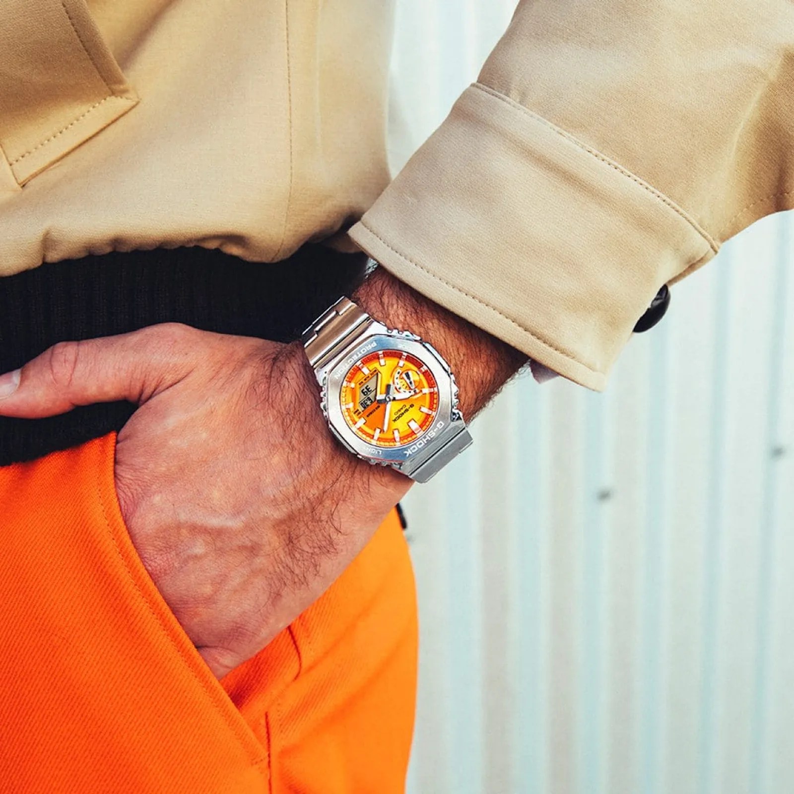 Silver metal G-Shock wristwatch with an orange face worn on a wrist over a beige jacket cuff and orange pants.